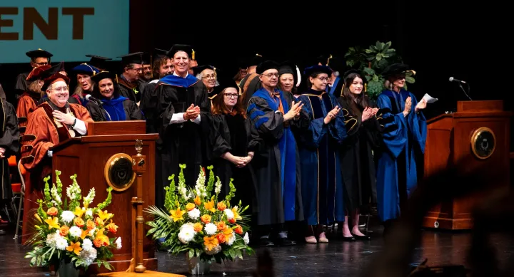 Faculty members in regalia stand and applaud at a commencement ceremony